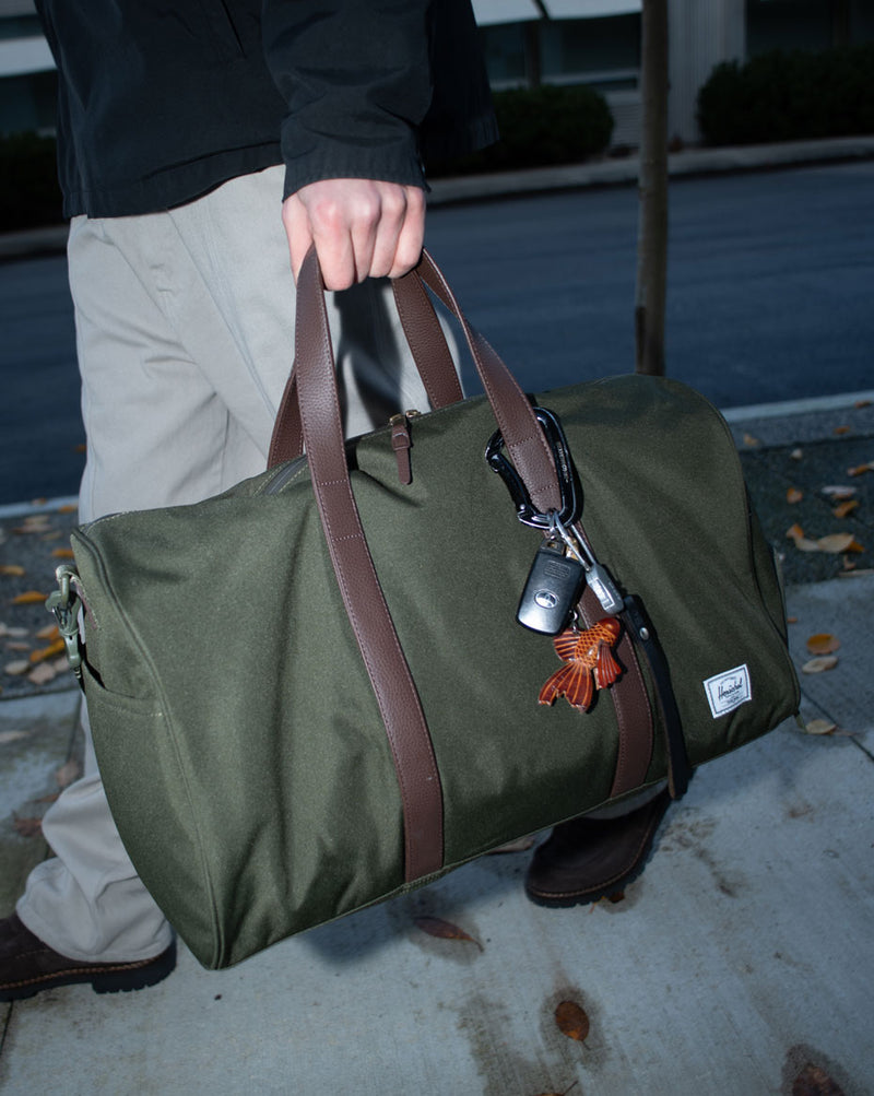 Herschel Supply Co. Novel Duffle bag in a multi-color camouflage pattern with black top handles and an adjustable shoulder strap.