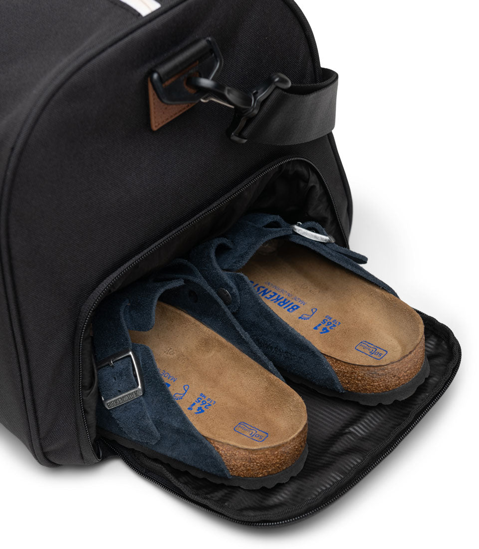Herschel Novel Duffle bag in a woodland camo pattern with black handles and a removable shoulder strap sitting on its pop-out stand.