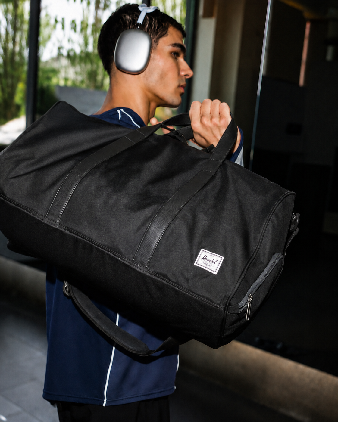 Angled view of a black Herschel Novel Duffle bag featuring a zip compartment for shoes, dual top handles, and a removable padded shoulder strap.