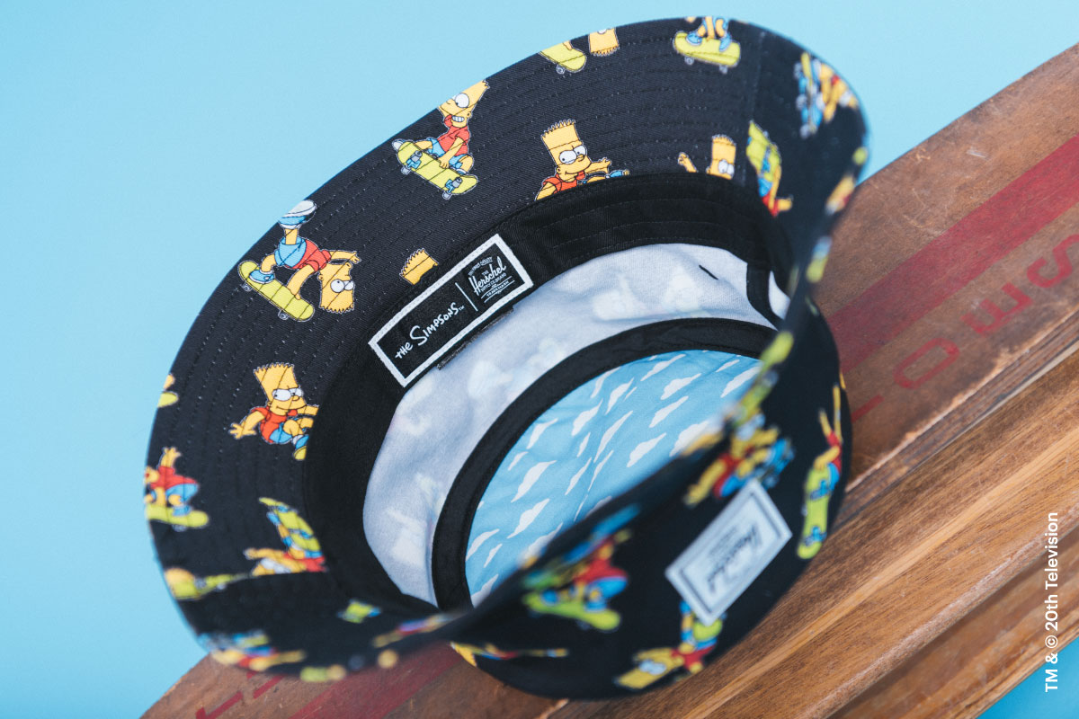 The inside cloud print on the Norman Bucket Hat in Bart Simpson print on a stack of skateboards with the Herschel Supply Company label on the front of the hat