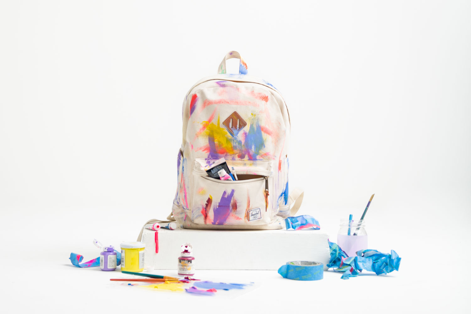 A Herschel Heavyweight Canvas Heritage Backpack in Natural covered in bright coloured paint next to a variety of painting utensils and paints