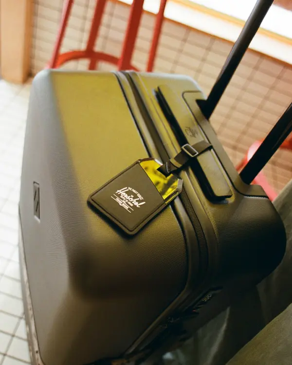 A close up photograph of a large black herschel heritage hardshell luggage with a luggage tag hanging over the zipper showing the herschel classic logo.